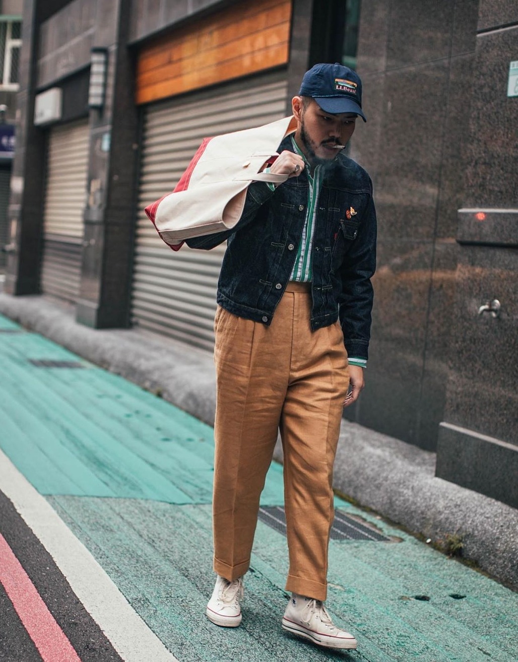 Le pantalon est en lin et dans une couleur décontractée (on s'éloigne du pantalon bleu marine ou du pantalon gris) mais il reste associé à un certain classicisme par sa coupe (taille haute, ample, présence de pinces et d'un bas revers). La présence d'une chemise décontractée permet de faire le lien avec la veste en jean, la tenue aurait été moins réussie avec une chemise en popeline bleu clair ou blanche par exemple. La tenue aurait très bien pu intégrer des derbies en lieu et place d'une paire de converse, cela n'aurait pas changé son ton.
Selon nous cette tenue doit simplement servir d'inspiration, en situation réelle il y a de fortes chances qu'elle fasse déguisement. Cet effet déguisement est dû à la grande disparité de couleurs, aux différents registres utilisés (veste en jean et pantalon en lin) mais aussi au fait que chaque pièce possède une identité assez forte.