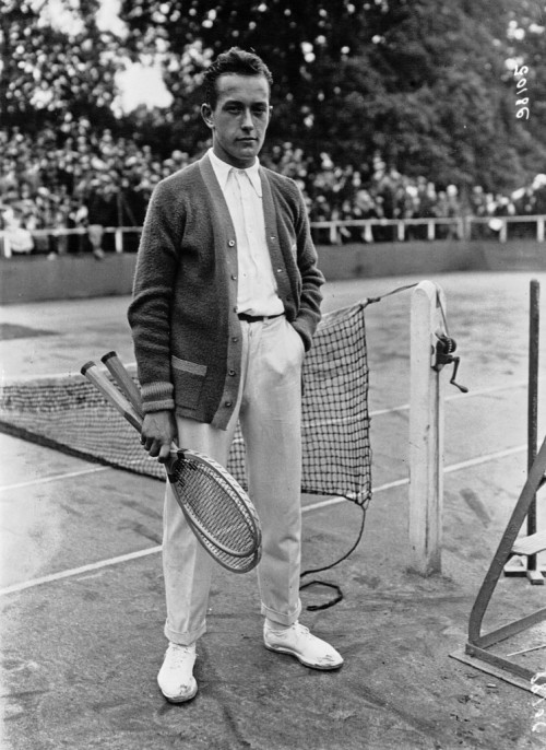 Henri Cochet en 1922, on remarque les sneakers portées avec un pantalon (en coton?) feu de plancher porté à la taille naturelle avec une chemise oxford et un cardigan. Le cardigan offre un effet de texture assez sympatique et qui est cohérent avec la tenue sportive. On remarque que bien que datée d’un siècle la tenue reste d’une surprenante actualité puisqu’on retrouve grosso modo la même construction dans la tenue montrée en début d’article (voir photo suivante).